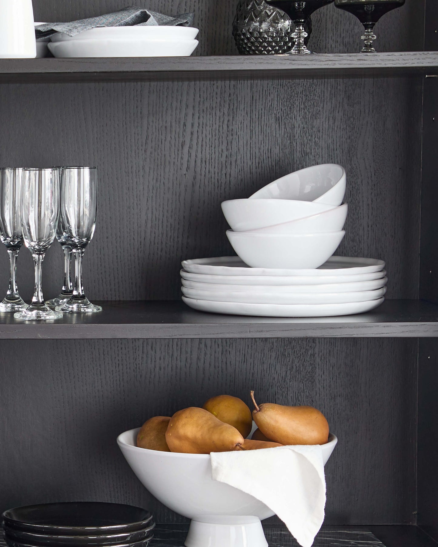 
                  
                    White Khira Dinnerware stacked on modern black hutch with champagne glasses and pedestal bowl.
                  
                