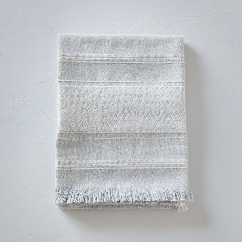 
                  
                    La Marea baby alpaca throw blanket folded with textured white stripes and light gray base for luxury gifts.
                  
                