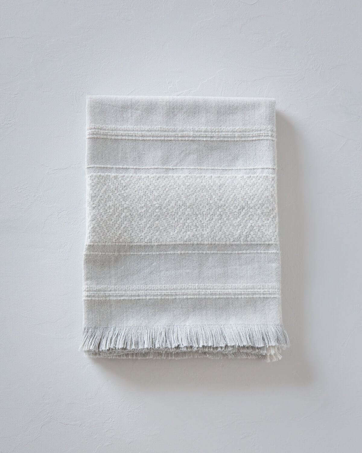 
                  
                    La Marea baby alpaca throw blanket folded with textured white stripes and light gray base for luxury gifts.
                  
                