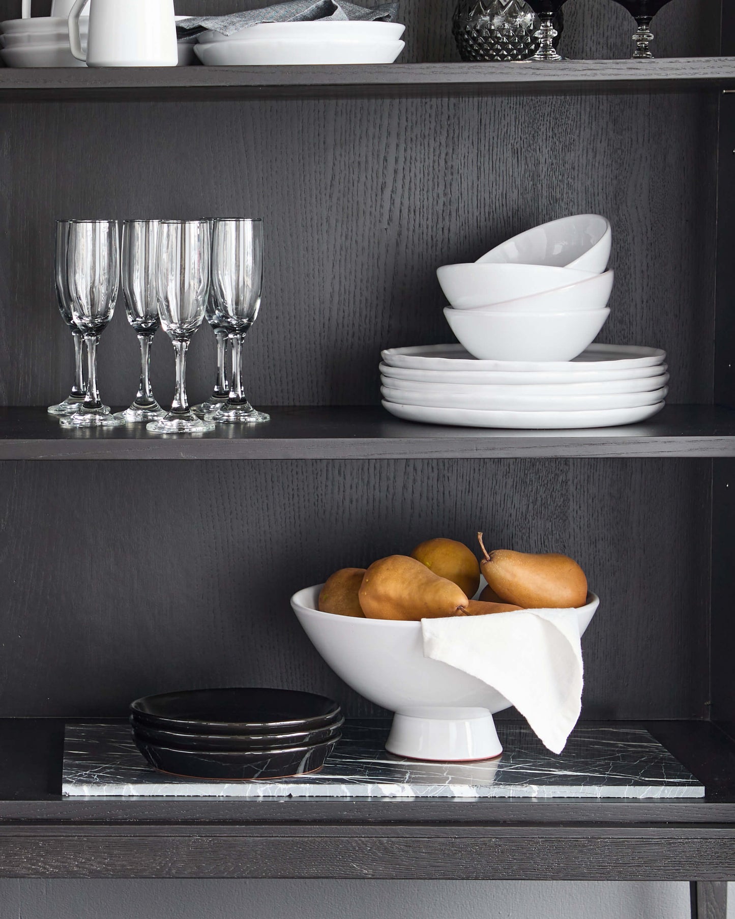 
                  
                    Modern hutch with ceramic dinnerware and white pedestal bowl filled with golden pears.
                  
                