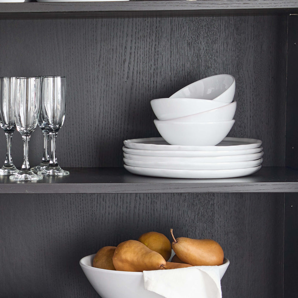 
                  
                    White Khira Dinnerware stacked on modern black hutch with champagne glasses and pedestal bowl.
                  
                