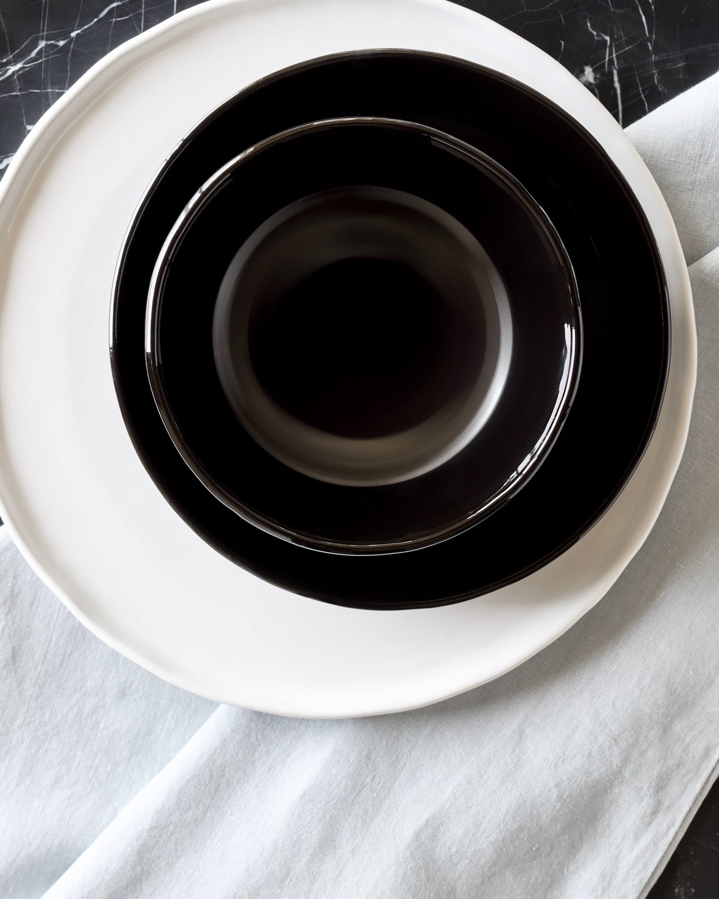 
                  
                    Glossy black ceramic dinnerware stacked on marble table with blue tablecloth. Modern, handcrafted dinnerware.
                  
                